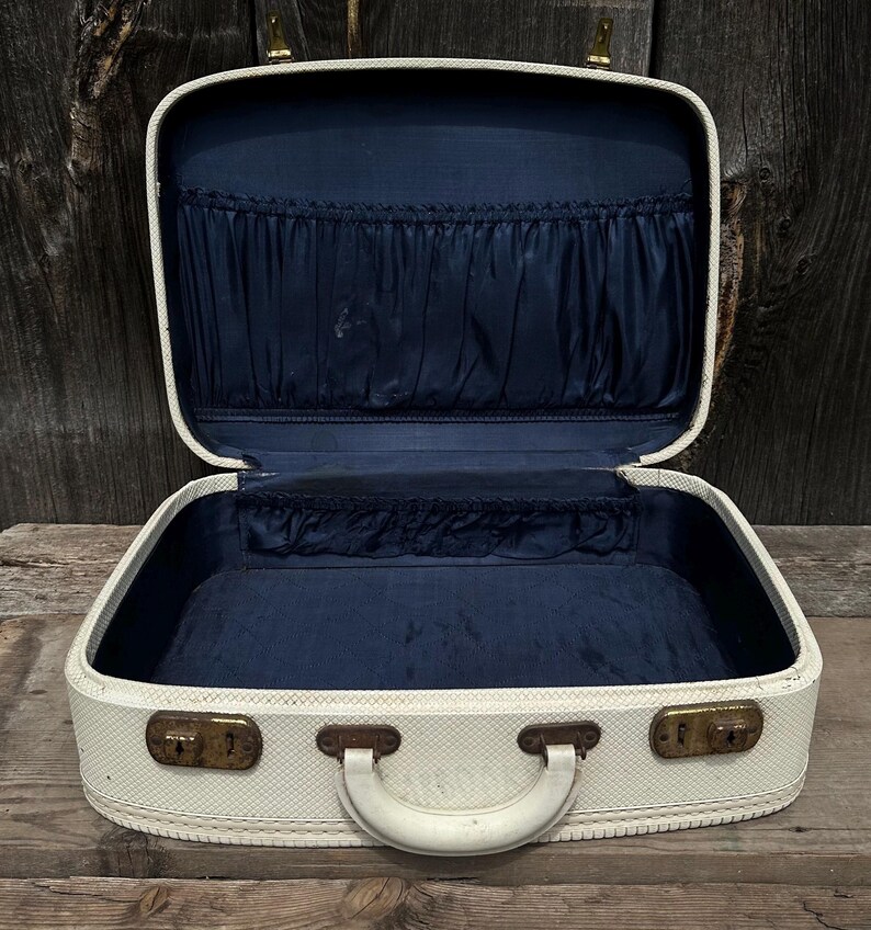 May include: Open vintage cream-colored suitcase with a navy blue interior. The suitcase has a white handle, brass-colored latches, and a textured exterior. The interior features pockets and a lined base, suggesting it's designed for travel or storage.
