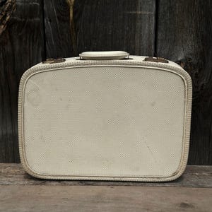 May include: Cream-colored vintage suitcase with a textured, cross-hatched pattern. Features a rounded rectangular shape, a top handle, and brass-toned hardware. The suitcase is set against a weathered wooden background.
