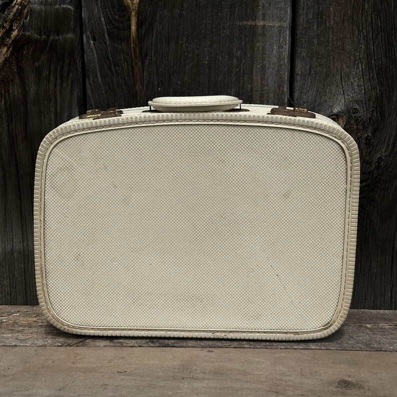 May include: Cream-colored vintage suitcase with a textured, cross-hatched pattern. Features a rounded rectangular shape, a top handle, and brass-toned hardware. The suitcase is set against a weathered wooden background.