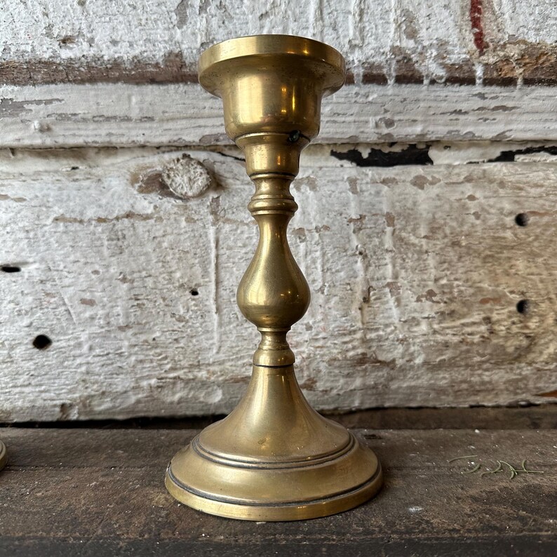 May include: A vintage brass candlestick holder with a classic design. The candlestick has a wide base, a slender, ornate stem, and a flared top to hold a candle. The brass has a warm, golden hue, and the piece appears to be aged.