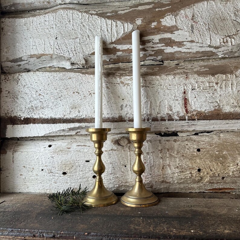 May include: Two brass candle holders with white candles. The candle holders have a classic design with a round base and a slender stem. A small sprig of greenery rests near the base of the candle holders. The background is a weathered, white-painted wooden surface.