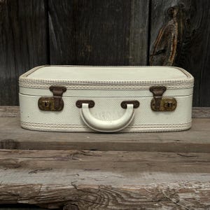 May include: A vintage off-white suitcase with a textured pattern, featuring a cream-colored handle and brass-toned clasps. The suitcase is rectangular with rounded edges and sits on a wooden surface, evoking a sense of travel and nostalgia.