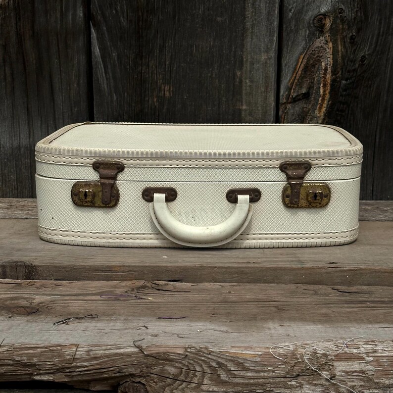 May include: A vintage off-white suitcase with a textured pattern, featuring a cream-colored handle and brass-toned clasps. The suitcase is rectangular with rounded edges and sits on a wooden surface, evoking a sense of travel and nostalgia.