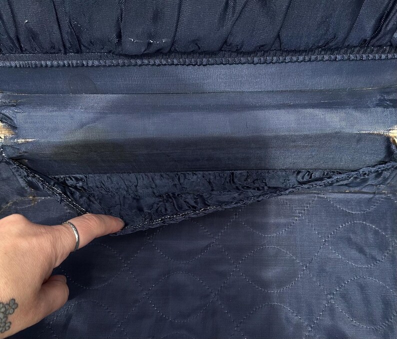 May include: Close-up of a vintage navy blue suitcase interior. The interior features a quilted pattern on the bottom section and a pleated fabric detail at the top. The lining shows signs of wear and age.