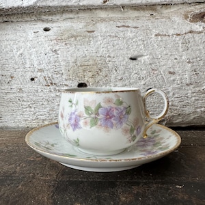 Könnte beinhalten: Weißes Tee-Set mit Tasse und Untertasse, verziert mit lila, rosa und grünen Blumenmustern. Tasse und Untertasse sind mit Gold verziert. Das Set steht auf einer dunklen Holzoberfläche.