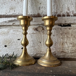 May include: Two antique brass candlesticks with white candles. The candlesticks have a classic design with a round base, a slender stem, and a flared top. The brass has a warm, aged patina, and the candles are unlit.