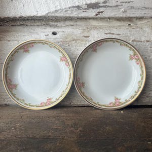 May include: Two white ceramic bowls with a pink rose and gold trim design. The bowls are round and have a shallow depth. The floral pattern is repeated around the rim of each bowl. The bowls are set on a wooden surface.