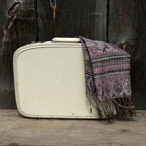 Könnte beinhalten: Ein cremefarbener Vintage-Koffer mit strukturierter Oberfläche und einem dekorativen Schal, der darüber drapiert ist. Der Schal hat ein Paisley-Muster in Lila-, Braun- und Grautönen mit Fransen.