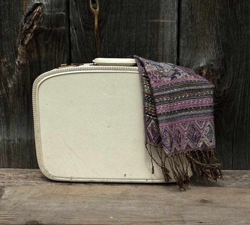 May include: A vintage cream-colored suitcase with a textured surface and a decorative scarf draped over it. The scarf features a paisley pattern in shades of purple, brown, and gray, with fringe.