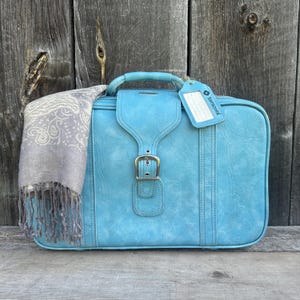 Op de afbeelding: Een lichtblauwe rechthoekige koffer met een centrale gespriem en een handvat aan de bovenkant. Een grijze sjaal met patroon en franjes hangt over de linkerkant. Een blauw bagagelabel met de tekst Samsonite is aan het handvat bevestigd.