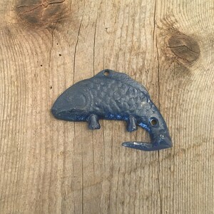 May include: A dark blue metal fish-shaped hook with a textured surface. The hook is attached to a wooden surface.