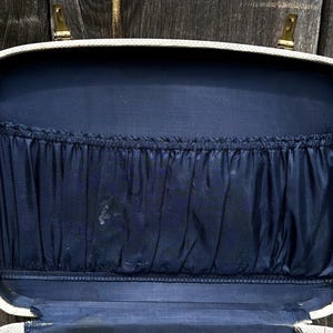 May include: Open vintage suitcase with a cream-colored exterior and a navy blue interior. The interior features a pleated pocket. The suitcase has gold-colored latches and hinges. The background is a weathered wooden surface.