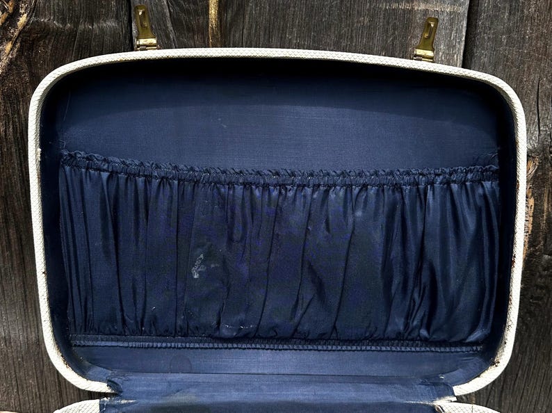May include: Open vintage suitcase with a cream-colored exterior and a navy blue interior. The interior features a pleated pocket. The suitcase has gold-colored latches and hinges. The background is a weathered wooden surface.
