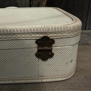 May include: Off-white vintage suitcase with a textured, diamond-patterned exterior. The suitcase features a decorative woven trim and a dark metal clasp. The rounded edges and overall design suggest a classic, travel-themed aesthetic.