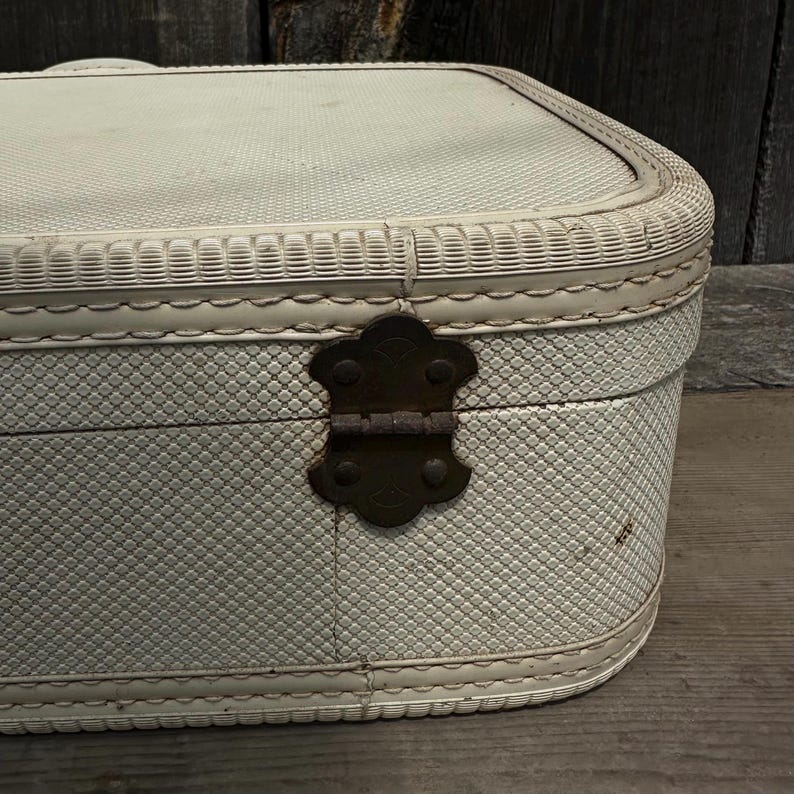 May include: Off-white vintage suitcase with a textured, diamond-patterned exterior. The suitcase features a decorative woven trim and a dark metal clasp. The rounded edges and overall design suggest a classic, travel-themed aesthetic.