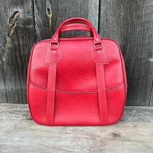 May include: A vintage red bowling bag with a top handle and two vertical straps. The bag has a rectangular shape with rounded corners and a textured surface. The bag is made of a durable material and is designed to carry bowling balls and accessories.