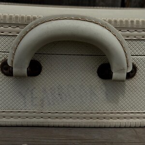 May include: Close-up of a vintage suitcase with a cream-colored handle and textured exterior. The handle is stitched with a contrasting thread and attached with dark brown metal hardware. The suitcase has a diamond pattern.