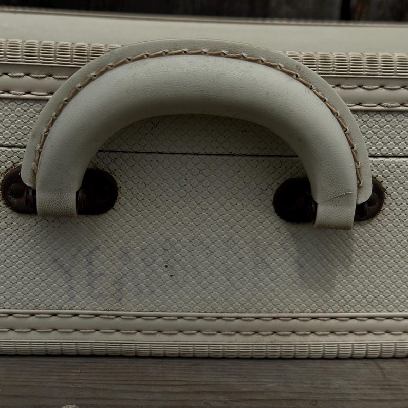 May include: Close-up of a vintage suitcase with a cream-colored handle and textured exterior. The handle is stitched with a contrasting thread and attached with dark brown metal hardware. The suitcase has a diamond pattern.