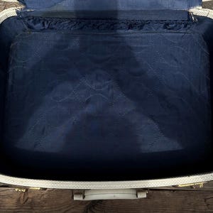 May include: An open, vintage suitcase with a white exterior and a navy blue interior. The interior is quilted with a repeating pattern. The suitcase has gold-colored hardware and a white handle. The suitcase is sitting on a wooden surface.