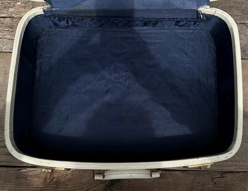 May include: An open, vintage suitcase with a white exterior and a navy blue interior. The interior is quilted with a repeating pattern. The suitcase has gold-colored hardware and a white handle. The suitcase is sitting on a wooden surface.