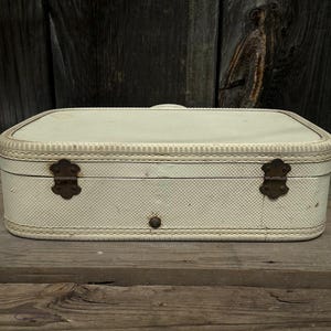 May include: Cream-colored vintage suitcase with a textured pattern and decorative stitching. Features two ornate metal clasps and a central button closure. The suitcase is rectangular with rounded corners.
