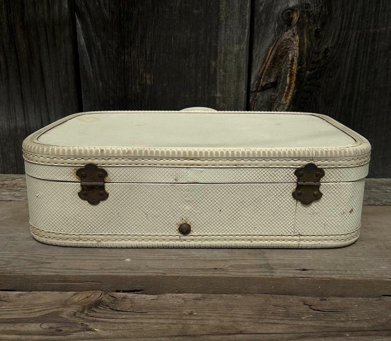 May include: Cream-colored vintage suitcase with a textured pattern and decorative stitching. Features two ornate metal clasps and a central button closure. The suitcase is rectangular with rounded corners.
