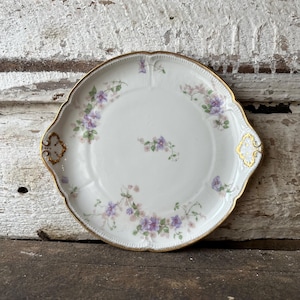 May include: White porcelain serving plate with a scalloped edge and gold trim. The plate is decorated with delicate purple and green floral patterns. Two gold handles are on either side of the plate.