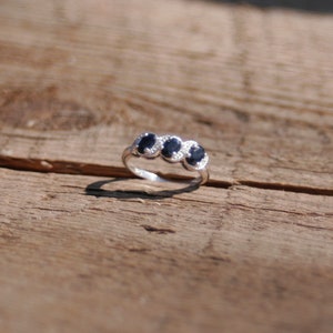May include: A silver ring featuring three oval-shaped black gemstones set in a row. Each gemstone is surrounded by a halo of smaller white gemstones.