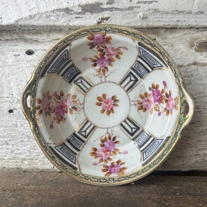 May include: A vintage porcelain dish with a floral design. The dish features pink roses, gold trim, and geometric black and white patterns. It has two handles and a scalloped edge, showcasing a classic aesthetic.