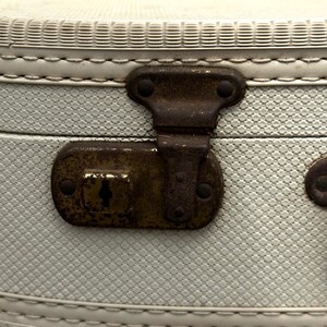 May include: Close-up of an antique suitcase latch. The latch is made of aged, rusty metal and is set against a light blue textured background. The suitcase has a white stitched trim.