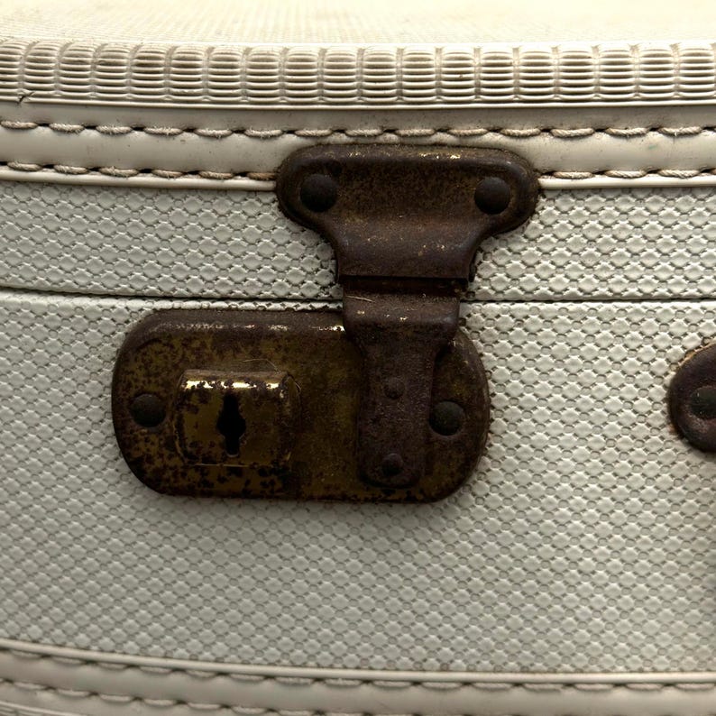 May include: Close-up of an antique suitcase latch. The latch is made of aged, rusty metal and is set against a light blue textured background. The suitcase has a white stitched trim.