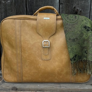 May include: A vintage Samsonite suitcase in a warm tan color, featuring a top handle, buckle closure, and a decorative stitched design. A green paisley scarf with fringe is draped over the suitcase. The suitcase is on a wooden surface.