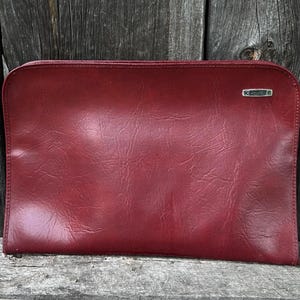 May include: A rectangular dark red document case with a smooth, slightly textured surface and rounded corners. A silver-toned rectangular metal plaque is attached to the upper right corner, featuring small, illegible embossed text.