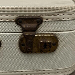 May include: Close-up of a vintage, light blue suitcase with a textured pattern. The suitcase features a tarnished, antique brass clasp and hinges. The edges are trimmed with white stitching and a decorative border.