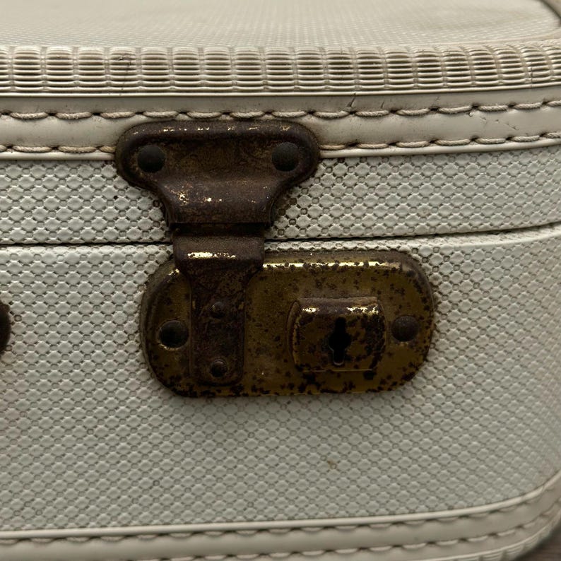 May include: Close-up of a vintage, light blue suitcase with a textured pattern. The suitcase features a tarnished, antique brass clasp and hinges. The edges are trimmed with white stitching and a decorative border.