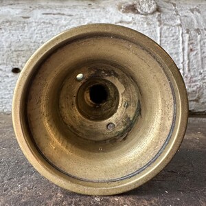 May include: A close-up view of a vintage brass candlestick holder. The holder has a circular base with a central well and two small holes. The brass has a warm, aged patina, with darker areas suggesting age and use.