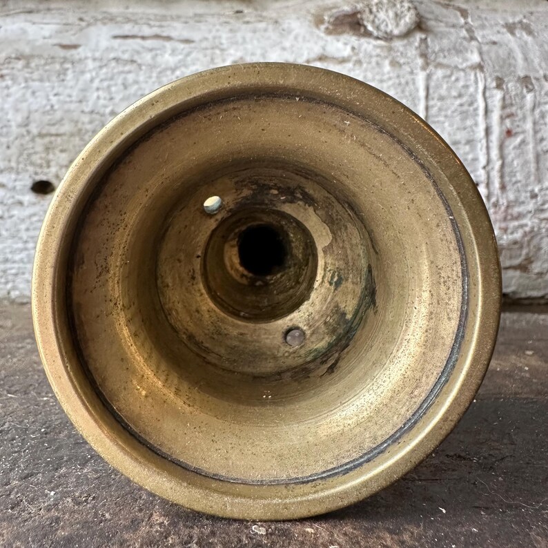 May include: A close-up view of a vintage brass candlestick holder. The holder has a circular base with a central well and two small holes. The brass has a warm, aged patina, with darker areas suggesting age and use.