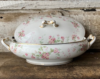 May include: A white porcelain serving dish with a lid, featuring a delicate floral pattern of pink and green flowers. The dish has a gold trim around the edges and a gold handle on the lid.