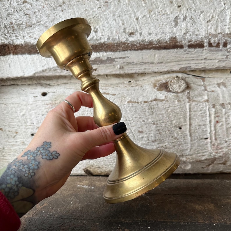 May include: A vintage brass candlestick holder with a wide base and a flared top. The candlestick has a bulbous center and a smooth, polished finish. The brass has a warm, golden hue, and the design is classic and elegant.