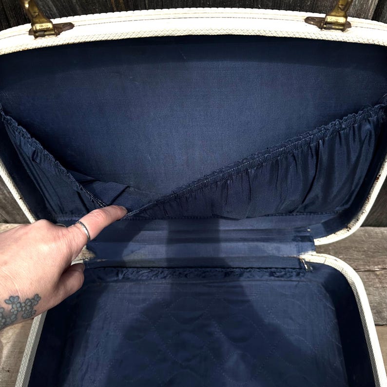 May include: Open vintage white suitcase with brass hardware and a navy blue interior. The interior features pockets and a quilted bottom. The suitcase is open, revealing its storage space.
