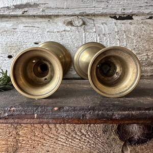 May include: Four antique brass candle holders. The candle holders have a round base and a flared, cup-like top. The brass has a warm, aged patina. The background is a weathered, white-painted wooden surface.