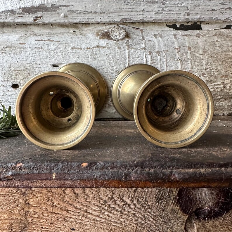 May include: Four antique brass candle holders. The candle holders have a round base and a flared, cup-like top. The brass has a warm, aged patina. The background is a weathered, white-painted wooden surface.