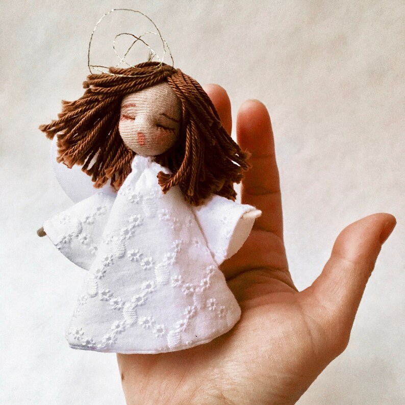 May include: A small white fabric angel doll with brown yarn hair and a halo. The doll is wearing a white dress with a floral pattern and has closed eyes.