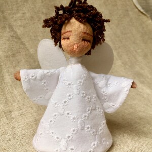 May include: A white fabric angel doll with brown curly hair and closed eyes. The doll is wearing a white dress with a lace pattern and has white wings.