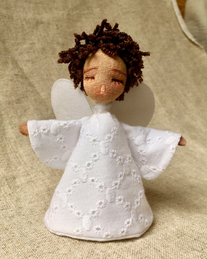 May include: A white fabric angel doll with brown curly hair and closed eyes. The doll is wearing a white dress with a lace pattern and has white wings.
