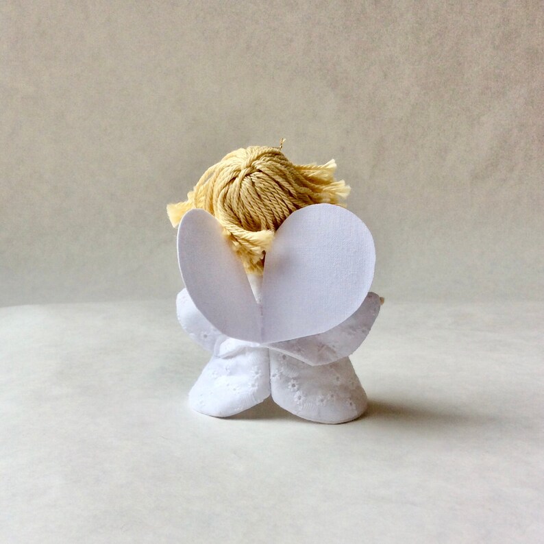 May include: A white angel figurine with paper wings and blonde yarn hair. The angel is facing away from the camera.