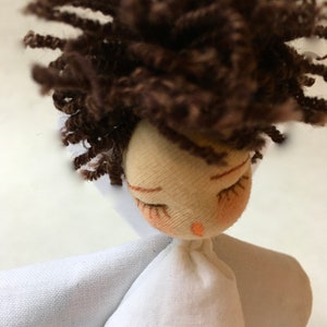 May include: A handmade doll with brown curly hair, closed eyes, and a white dress with a flower embroidered on the front.