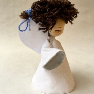 May include: A handmade doll with brown yarn hair, closed eyes, and a white fabric dress with a blue sleeve. The doll is holding a white paper wing.