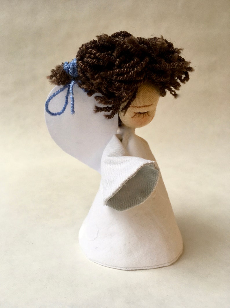 May include: A handmade doll with brown yarn hair, closed eyes, and a white fabric dress with a blue sleeve. The doll is holding a white paper wing.