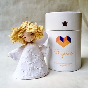 May include: A white fabric angel ornament with a blonde yarn hair and a white lace dress. The angel is packaged in a white cylinder box with a star and a heart logo that says "Ragskies" and includes the website address "www.etsy.com/shop/ragaskies" and the social media handle "@ragaskies".